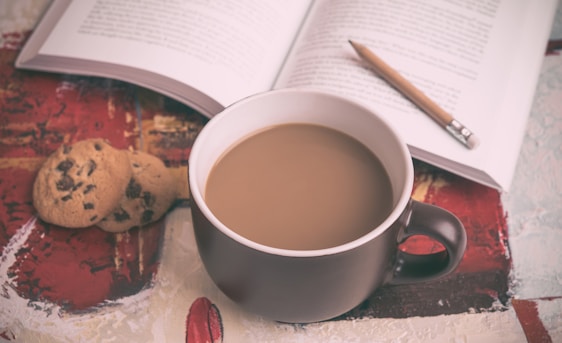 A cozy scene with a steaming cup of coffee in a dark mug placed on a surface with abstract patterns. Nearby are two chocolate chip cookies placed beside an open book. A pencil rests between the pages, suggesting a moment of relaxation and thought.