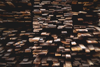 Stacks of wooden planks are arranged in a dense, layered formation. The varying shades of brown create a rustic texture and appearance, with some planks appearing freshly cut while others look aged. The angles and alignment of the planks form a geometric pattern that draws the eye through the image.