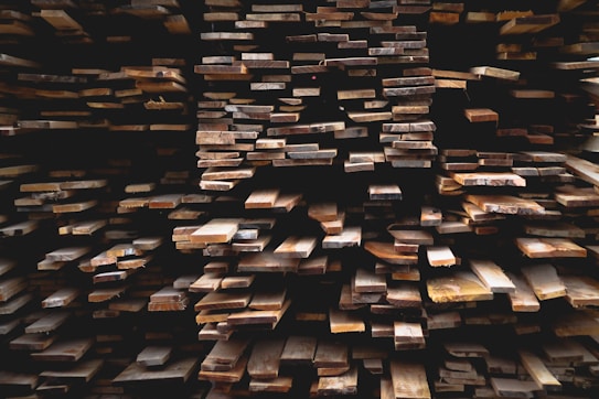 Stacks of wooden planks are arranged in a dense, layered formation. The varying shades of brown create a rustic texture and appearance, with some planks appearing freshly cut while others look aged. The angles and alignment of the planks form a geometric pattern that draws the eye through the image.