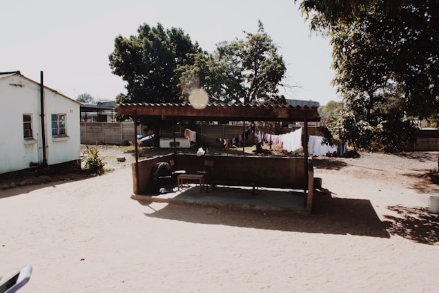 A rural setting with a small, simple shelter made of wood and corrugated metal situated in a sandy open area. Nearby, a modest house with a white exterior and blue window frames stands beside a dirt patio. Clothes are hung on lines to dry in the background, creating a sense of daily life activities. Tall trees provide some shade, casting soft shadows on the ground.