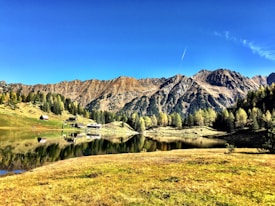 A serene mountainous landscape with a clear blue sky above. Rugged, rocky mountains form the backdrop, with a mix of evergreen and deciduous trees in the foreground. A small, calm lake reflects the surrounding scenery perfectly. A few rustic cabins dot the landscape, adding a touch of human presence to the natural setting.