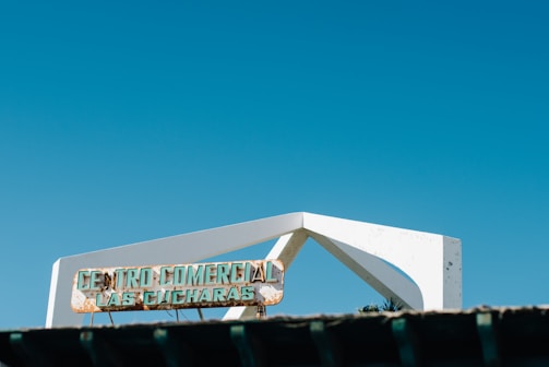 A sign reads 'Centro Comercial Las Cucharas,' mounted on a white angular structural element against a clear blue sky. The sign appears weathered and partially corroded.