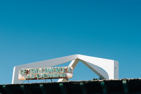 A sign reads 'Centro Comercial Las Cucharas,' mounted on a white angular structural element against a clear blue sky. The sign appears weathered and partially corroded.
