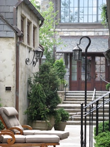 A cozy and elegantly designed courtyard features stone buildings with charming wrought iron light fixtures. Lush green potted plants and trees line the pathway leading to stone steps that ascend to a set of glass doors framed by large windows. A comfortable cushioned lounge chair is placed on the stone patio, enhancing the serene atmosphere.