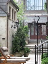 A cozy and elegantly designed courtyard features stone buildings with charming wrought iron light fixtures. Lush green potted plants and trees line the pathway leading to stone steps that ascend to a set of glass doors framed by large windows. A comfortable cushioned lounge chair is placed on the stone patio, enhancing the serene atmosphere.