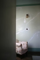 A vintage armchair with a floral pattern is placed against a wall with a faded green hue. Above, a small vintage light fixture is mounted on the wall. The room appears dimly lit, with natural light casting subtle shadows.