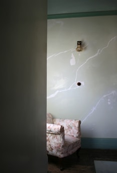 A vintage armchair with a floral pattern is placed against a wall with a faded green hue. Above, a small vintage light fixture is mounted on the wall. The room appears dimly lit, with natural light casting subtle shadows.