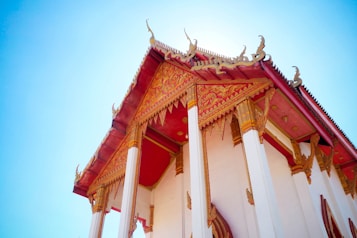 A close-up view of a temple with ornate, intricate designs. The roof is detailed with gold embellishments against a rich maroon background. The pillars are white with golden accents, reflecting traditional architectural styles. The temple is set against a clear blue sky, enhancing the vibrant colors.