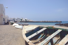 An old, weathered boat resting on a sandy shore with a row of small, white boats lined up next to it. The background features a calm, turquoise sea and a long breakwater stretching into the water. White buildings with flat roofs and green shutters are visible along the coast under a clear blue sky.