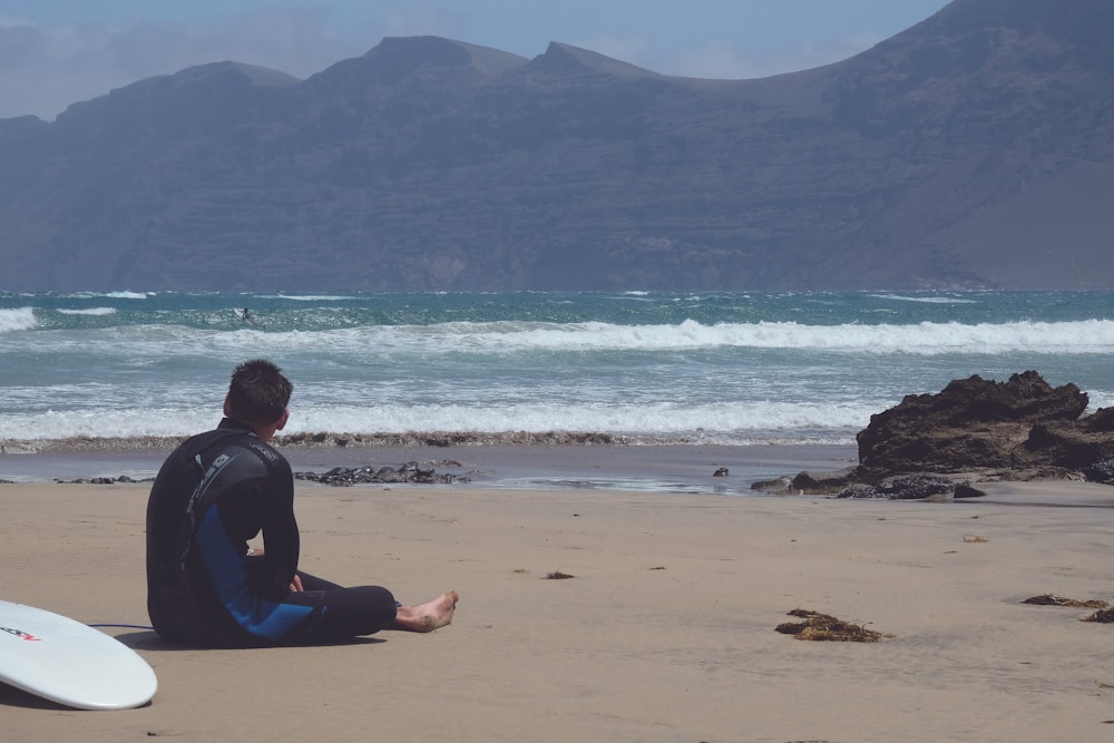 man in black shirt and blue pants sitting on beach shore during daytime