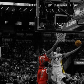 A basketball game scene with a player in a white jersey performing a layup as a player in a red jersey attempts to block the shot. The action is intense, with a crowded stadium visible in the background. The image is predominantly black and white, except for the red jerseys and the yellow and orange hues of the ball and part of the player's attire.