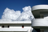 A modern building with curved balconies and a flat rooftop is set against a backdrop of large, fluffy clouds in a clear blue sky.