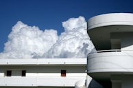 A modern building with curved balconies and a flat rooftop is set against a backdrop of large, fluffy clouds in a clear blue sky.
