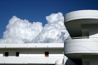 A modern building with curved balconies and a flat rooftop is set against a backdrop of large, fluffy clouds in a clear blue sky.