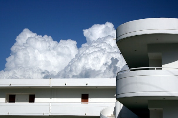 A modern building with curved balconies and a flat rooftop is set against a backdrop of large, fluffy clouds in a clear blue sky.