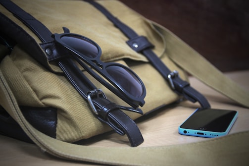 A close-up of a khaki-colored bag with black leather straps resting on a wooden surface. A pair of black sunglasses is placed on top of the bag, and a smartphone with a blue case lies next to it.