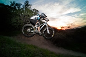 A person is riding a mountain bike downhill on a trail surrounded by greenery. The sun is setting in the background, creating a dramatic sky with hues of orange and blue. The cyclist is wearing a helmet and protective gear.