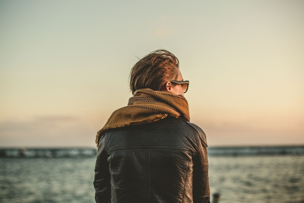 Fokusfoto einer Person, die tagsüber eine schwarze Lederjacke mit braunem Schal in der Nähe des Meeres trägt