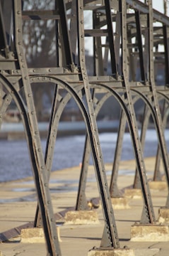 Metallic frame structure with rivets, positioned along a concrete waterfront. The blurred background features water and trees, hinting at an outdoor setting possibly along a river or canal.