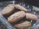 A glass bowl containing several brown cookies stacked on each other. The cookies appear freshly baked and have a coarse texture with visible chocolate pieces.
