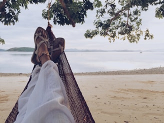 A person relaxes on a hammock facing a serene beach, with their legs stretched out and sandals on. The scene is framed by overhanging tree branches, with a view of calm waters and distant islands. The sky is overcast, creating a peaceful and tranquil atmosphere.