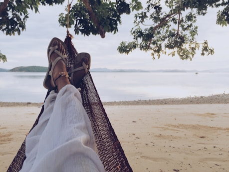 A person relaxes on a hammock facing a serene beach, with their legs stretched out and sandals on. The scene is framed by overhanging tree branches, with a view of calm waters and distant islands. The sky is overcast, creating a peaceful and tranquil atmosphere.