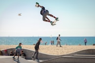 A skateboarder is performing an aerial trick against the backdrop of a beach with the ocean in the distance. The skateboarder is in mid-air, showcasing impressive skill and balance. Below, other people, including a child also on a skateboard, are enjoying the skate park. The sandy beach and a calm sea with sailboats add a serene atmosphere to the active scene.