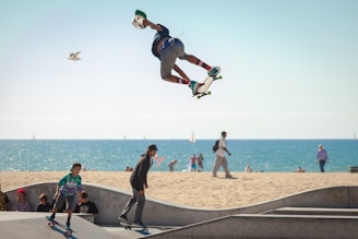 A skateboarder is performing an aerial trick against the backdrop of a beach with the ocean in the distance. The skateboarder is in mid-air, showcasing impressive skill and balance. Below, other people, including a child also on a skateboard, are enjoying the skate park. The sandy beach and a calm sea with sailboats add a serene atmosphere to the active scene.