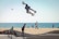A skateboarder is performing an aerial trick against the backdrop of a beach with the ocean in the distance. The skateboarder is in mid-air, showcasing impressive skill and balance. Below, other people, including a child also on a skateboard, are enjoying the skate park. The sandy beach and a calm sea with sailboats add a serene atmosphere to the active scene.