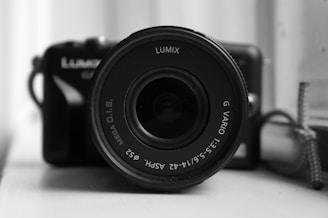 A close-up view of a camera lens, prominently displaying brand and lens specifications. The background shows parts of the camera body, slightly out of focus. Monochrome tone enhances the focus on the lens details.