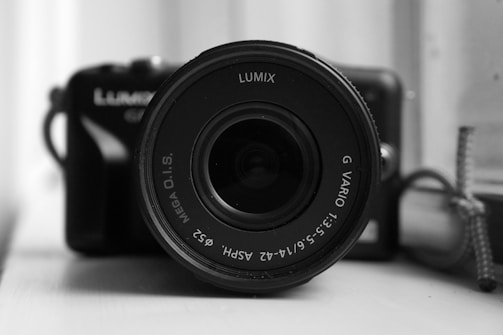 A close-up view of a camera lens, prominently displaying brand and lens specifications. The background shows parts of the camera body, slightly out of focus. Monochrome tone enhances the focus on the lens details.