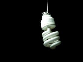A spiral-shaped compact fluorescent light bulb hanging from a white cord against a black background. The bulb is prominently featured and appears to be the only source of illumination in the scene.