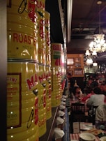 A row of large yellow cans labeled with tomatoes is aligned on a shelf in a dimly lit restaurant. In the background, diners are seated at tables with checkered tablecloths, contributing to a cozy and bustling atmosphere. The decor includes chandeliers and framed pictures on the walls.