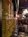 A row of large yellow cans labeled with tomatoes is aligned on a shelf in a dimly lit restaurant. In the background, diners are seated at tables with checkered tablecloths, contributing to a cozy and bustling atmosphere. The decor includes chandeliers and framed pictures on the walls.