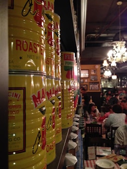 A row of large yellow cans labeled with tomatoes is aligned on a shelf in a dimly lit restaurant. In the background, diners are seated at tables with checkered tablecloths, contributing to a cozy and bustling atmosphere. The decor includes chandeliers and framed pictures on the walls.