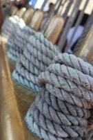 Thick ropes are neatly coiled around metallic cylindrical posts, suggesting a nautical setting. The ropes are light gray with hints of green, set against a blurred background with vertical lines and subtle warm tones.