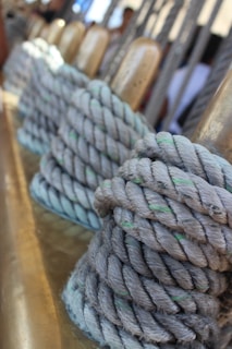 Thick ropes are neatly coiled around metallic cylindrical posts, suggesting a nautical setting. The ropes are light gray with hints of green, set against a blurred background with vertical lines and subtle warm tones.