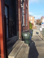 A sidewalk runs alongside a brick building with a series of large, green trash bins lined up against it. There are concrete steps leading to a dark, worn door. A street extends into the distance, with a stop sign and a few more brick buildings visible. The lighting suggests a sunny day with clear skies.
