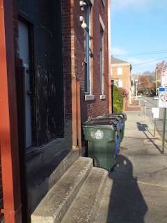 A sidewalk runs alongside a brick building with a series of large, green trash bins lined up against it. There are concrete steps leading to a dark, worn door. A street extends into the distance, with a stop sign and a few more brick buildings visible. The lighting suggests a sunny day with clear skies.