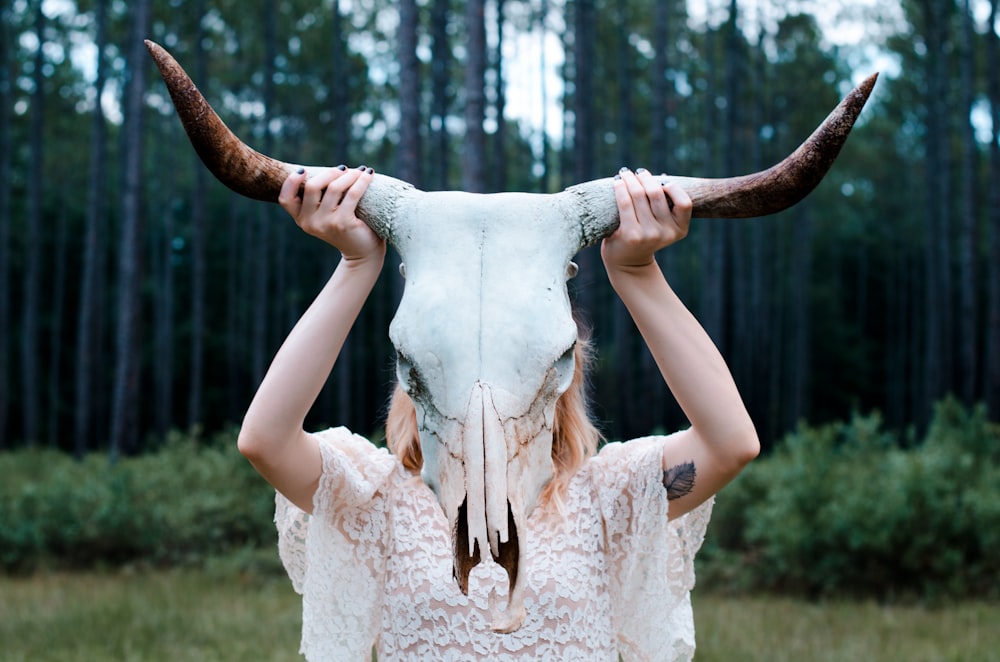 woman holding white and black animal skull wearing white lace floral sleeveless dress near green leaf trees