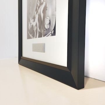 A black-framed picture rests on a surface with a black and white photograph depicting various mechanical parts inside. The frame is angled, showing its sleek, modern design, while the surface has a light, neutral tone.
