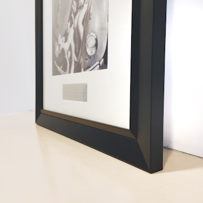 A black-framed picture rests on a surface with a black and white photograph depicting various mechanical parts inside. The frame is angled, showing its sleek, modern design, while the surface has a light, neutral tone.