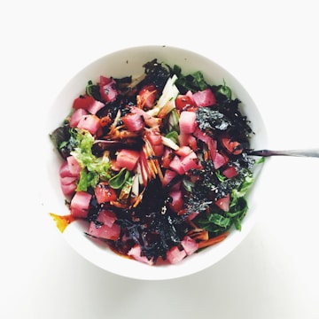 A vibrant salad with fresh greens, diced pink tuna, a mix of vegetables, and dark seaweed, drizzled with a dressing and served in a white bowl.