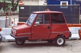 A compact red car with a boxy design is parked beside construction barriers. The vehicle has a single door visible on the side, small wheels, and a simple, vintage appearance. The background features construction site elements with a mix of orange and white traffic barriers, a blue temporary structure, and some greenery.