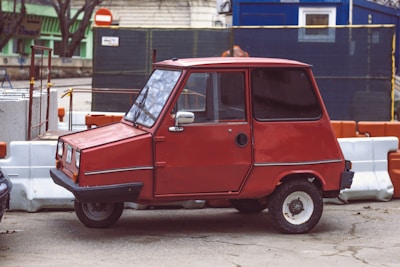 A compact red car with a boxy design is parked beside construction barriers. The vehicle has a single door visible on the side, small wheels, and a simple, vintage appearance. The background features construction site elements with a mix of orange and white traffic barriers, a blue temporary structure, and some greenery.