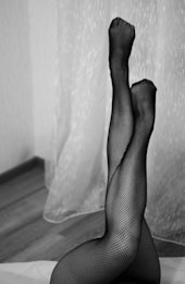 A pair of legs dressed in black fishnet stockings are raised and crossed at the ankles, positioned against a background of sheer drapes and a hardwood floor.