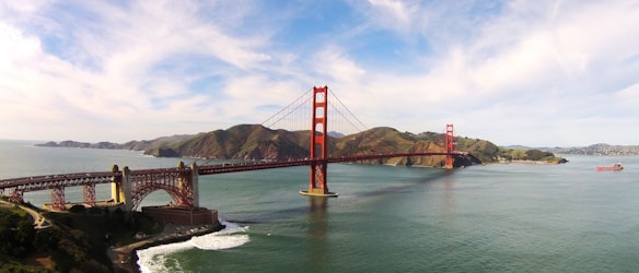 A large red suspension bridge spans across a body of water, with rolling hills and a partly cloudy sky in the background. A large cargo ship is seen in the water, and the shoreline includes rocky areas with light vegetation.