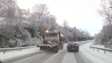 A snowplow with flashing lights drives down a snowy road, clearing the path. A car follows closely behind. Bare trees covered with snow line the sides of the road, creating a wintery landscape.