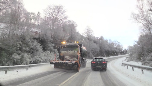 A snowplow with flashing lights drives down a snowy road, clearing the path. A car follows closely behind. Bare trees covered with snow line the sides of the road, creating a wintery landscape.