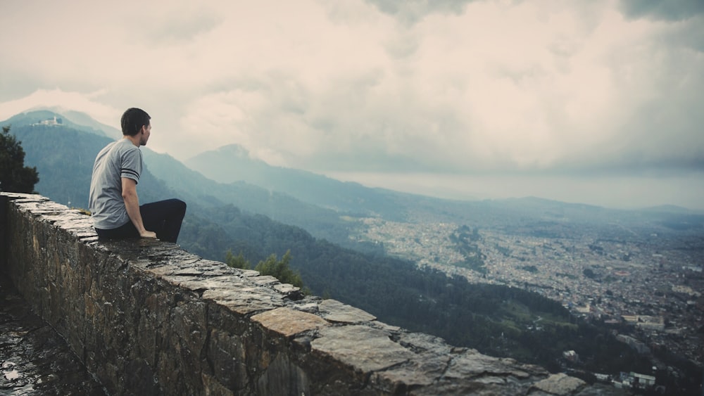 구름 낀 하늘 아래 산과 도시를 마주보고 있는 콘크리트 벽돌 벽에 혼자 앉아 있는 남자